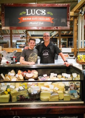 Two people smile below the Luc's sign and behind a fridge of cheeses