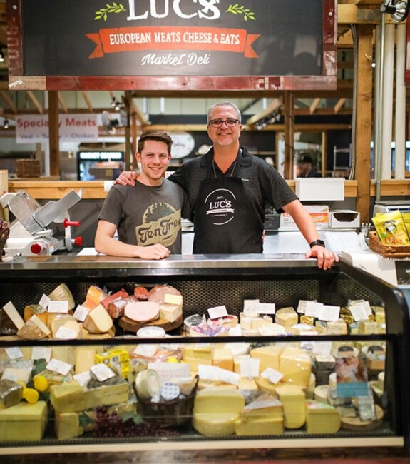 Two people smile below the Luc's sign and behind a fridge of cheeses
