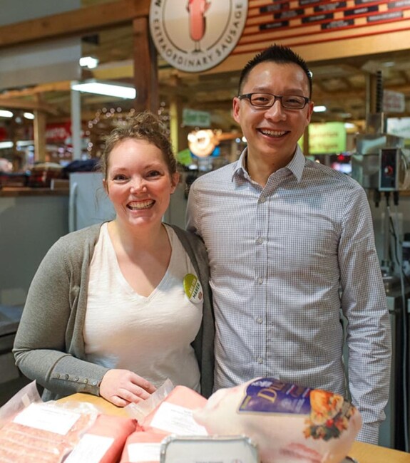Two people smiling behind a table of meat products