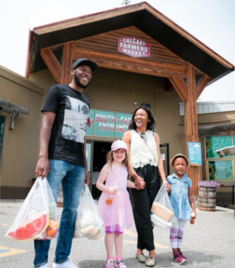 A family with two kids holding grocery bags stands outside the Market entrance