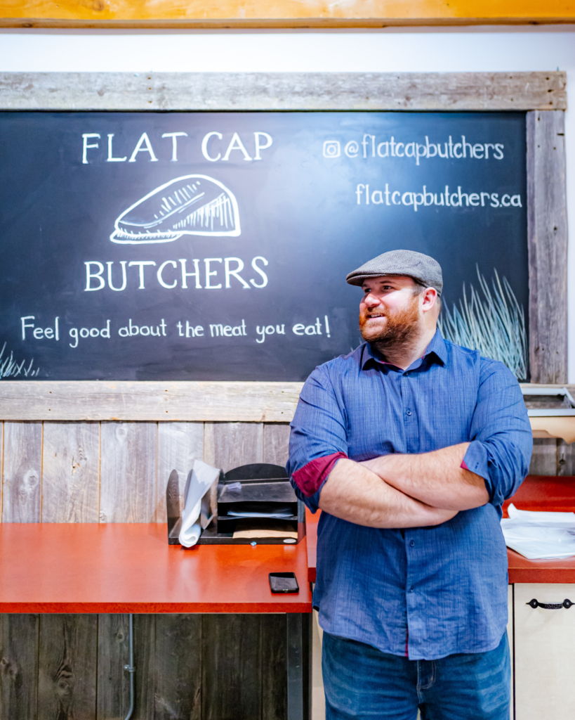 A person with crossed arms stands before the Flat Cap Butchers sign